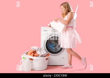 Fille en costume de fée avec bouteille de détergent à lessive près de la machine à laver et panier avec des vêtements sales sur fond rose Banque D'Images