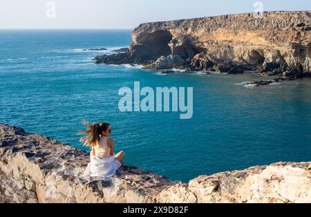 Une femme en robe blanche est assise sur un bord de falaise, surplombant l'océan bleu vaste et la côte rocheuse d'Ajuy sous le soleil brillant de midi. Banque D'Images