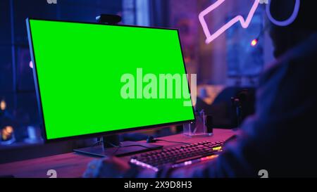 Joueur excité jouant à un jeu vidéo en ligne avec un écran vert maquette sur son puissant ordinateur de jeu personnel. Gros plan sur la pièce et le PC avec des lumières LED néon colorées. Soirée à la maison dans Loft Apartment. Banque D'Images