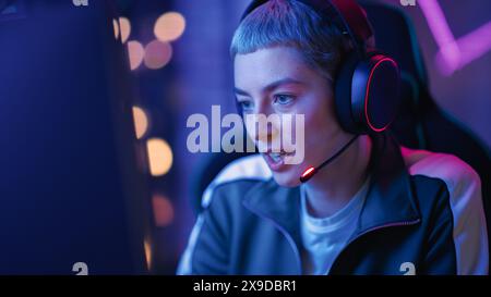 Joueur féminin professionnel jouant au jeu vidéo en ligne sur ordinateur. Gros plan Portrait de jeune femme élégante dans les écouteurs se battant dans le tournoi PvP avec d'autres joueurs, parler avec l'équipe sur microphone. Banque D'Images