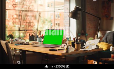 Ordinateur portable avec écran vert maquette écran Chroma Key debout sur le bureau dans l'espace de travail Loft moderne. Ouvrez Space Studio en arrière-plan avec vue fenêtre ville. Banque D'Images