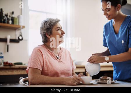 Soignant, sourire et femme avec du thé dans la cuisine de la maison de soins infirmiers pour le petit déjeuner, se détendre ou une nutrition saine de soutien. Infirmière, malade et nourriture pour sénior Banque D'Images