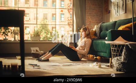 Jeune jolie femme aux cheveux blonds portant des écouteurs. Travailler à la maison sur ordinateur portable dans élégant Loft Apartment. Femme créative assise dans une posture détendue et vérifiant les médias sociaux. Banque D'Images