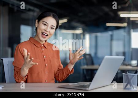 Femme asiatique joyeuse réussie sur le lieu de travail célébrant de bons résultats de réalisation élevée, femme d'affaires souriant et regardant la caméra, en utilisant un ordinateur portable au travail, femme gagnante satisfaite. Banque D'Images