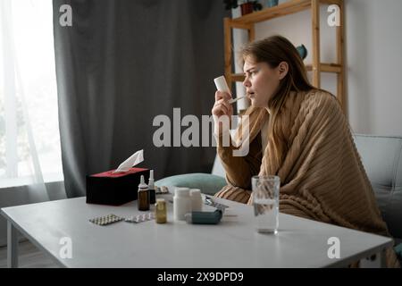 Vue de côté d'une jeune fille caucasienne malade assise à la maison sur le canapé, enveloppée dans une couverture, en utilisant un spray pour la gorge pour la toux. Le concept de traitement vi Banque D'Images