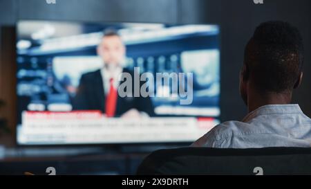 Man regarde Late Night News Show à la télévision tout en étant assis sur un canapé à la maison dans la soirée. Le présentateur parle à la télévision. Salon loft confortable et élégant avec lumières chaudes. Tir au-dessus de l'épaule. Banque D'Images