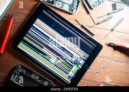 Tablette endommagée avec un écran déformé affichant des lignes colorées, entourée d'appareils électroniques démontés et d'outils de réparation sur une surface en bois Banque D'Images