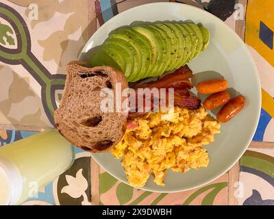 Vue aérienne d'une assiette de petit déjeuner avec œufs brouillés, tranches d'avocat, bacon, saucisses et pain, servi avec un verre de jus sur un t carrelé coloré Banque D'Images