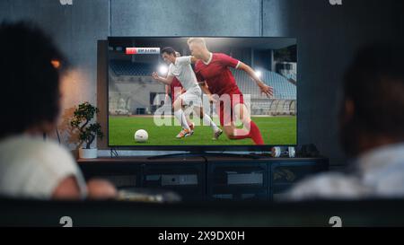 Couple noir regarde le match de football professionnel à la télévision, assis sur un canapé à la maison dans la soirée. Fans de football pour petit ami et petit ami regarder les sports. Vue arrière flou gros plan la nuit. Banque D'Images