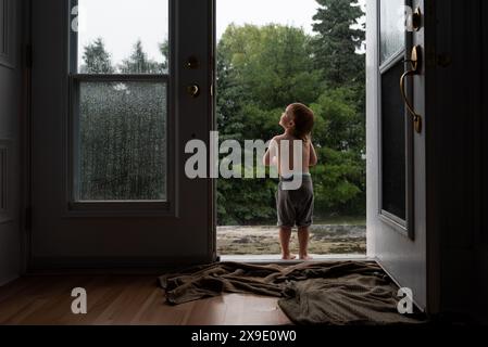 L'enfant se tient dans l'embrasure de la porte regardant la scène pluvieuse avec des arbres mouillés. Banque D'Images