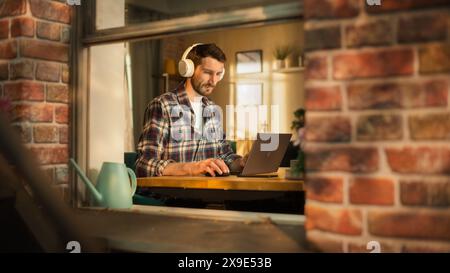 Bel homme caucasien travaillant sur un ordinateur portable tout en étant assis derrière son bureau au bureau à domicile. Entrepreneur indépendant écoutant de la musique, faisant du télétravail à distance. Vue de l'extérieur dans la fenêtre. Banque D'Images