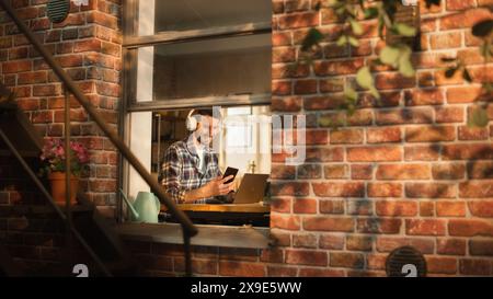 Freelancer moderne tapant sur un ordinateur portable et vérifie notification sur smartphone. Concept de professionnel e-Business entrepreneur travaillant à distance au bureau à domicile. Vue de l'extérieur dans la fenêtre. Banque D'Images