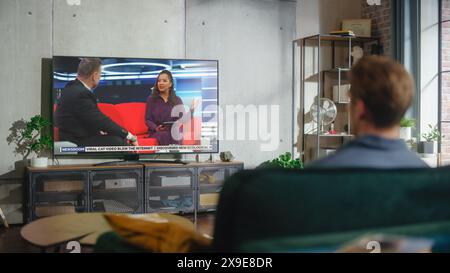 Jeune homme beau assis dans la pose détendue sur un canapé et regardant la télévision avec Live Talk Show pendant la journée le week-end. Sérieuse femme afro-américaine hôte interviewe invité dans le studio. Banque D'Images