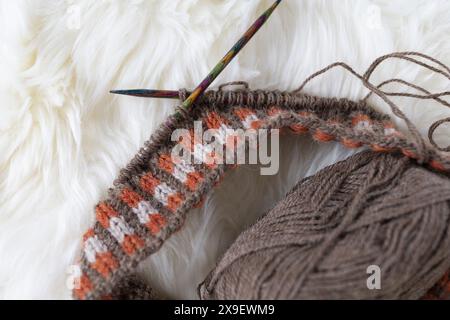 Photographie rapprochée de tricot Fair Isle en cours avec un projet de fil mince d'alpaga brun sur des aiguilles en bois colorées sur fourrure d'agneau blanche Banque D'Images