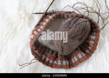 Photographie à plat de tricot Fair Isle en cours avec un projet de fil mince d'alpaga brun sur des aiguilles en bois colorées sur de la fourrure d'agneau blanche Banque D'Images