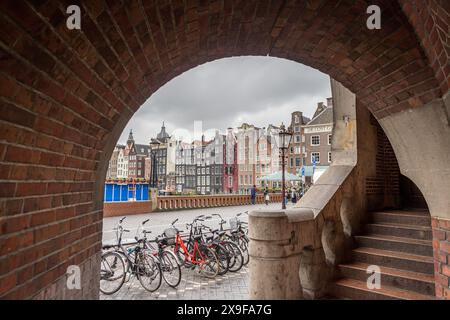 Une arche et une rangée de vélos stationnés encadrent les maisons hautes et inégales qui bordent Damrak à Amsterdam, aux pays-Bas vues le 29 mai 2024. Banque D'Images