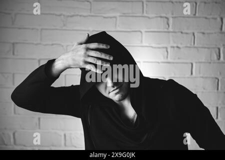 Un homme portant un sweat à capuche noir danse énergiquement avec des mouvements expressifs de la main devant un mur de briques blanches. Banque D'Images