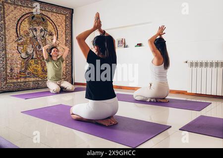 Groupe de jeunes dans un cours de yoga en position de prière avec les mains levées tout en étant assis sur le tapis à la salle de gym. Banque D'Images
