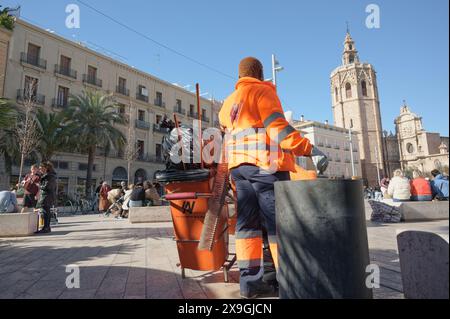 Un travailleur sanitaire en tenue orange vif nettoie les rues de Valence, avec des bâtiments historiques et une plaza de la reina animée en arrière-plan Banque D'Images