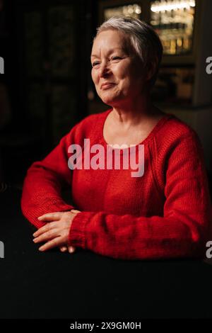 Portrait vertical d'une femme adulte mature joyeuse recevant une bonne prévision de l'avenir pendant la session de divination assise à table dans une pièce sombre, par la lumière Banque D'Images