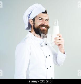 Beau chef barbu homme en uniforme avec bouteille de lait. Cuisine laitière. Cuisinier masculin souriant en uniforme avec une bouteille en verre de kéfir, yaourt, milkshake. Banque D'Images