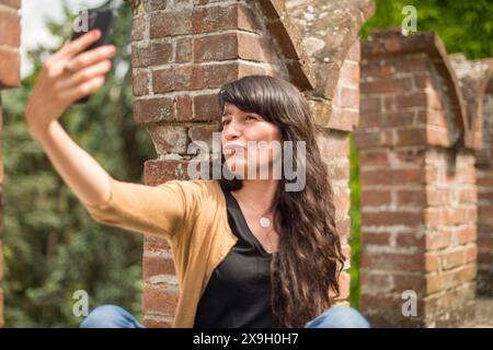 Jeune femme hispanique en vêtements décontractés axée sur la conversation avec smartphone dans un jardin paisible avec briques floues au premier plan Banque D'Images