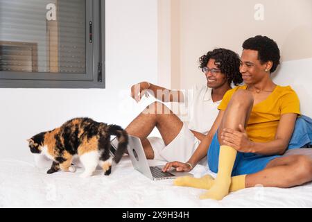 Couple gay utilisant un ordinateur portable dans la chambre à coucher à côté d'un chat Banque D'Images