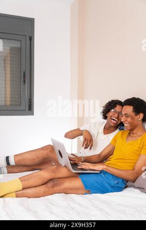 Photo verticale d'un couple gay latin heureux utilisant un ordinateur portable dans le lit Banque D'Images