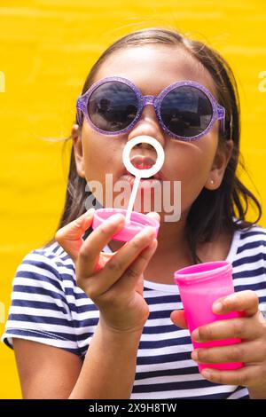 À l'école, une jeune fille biraciale portant des lunettes de soleil souffle des bulles à l'extérieur Banque D'Images