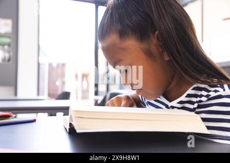 À l'école, jeune fille biraciale se concentrant sur la lecture d'un livre dans la classe Banque D'Images