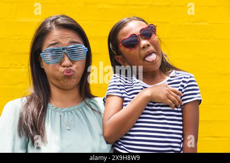 Devant un mur jaune à l'extérieur, une jeune fille et une femme biraciales font des visages drôles Banque D'Images