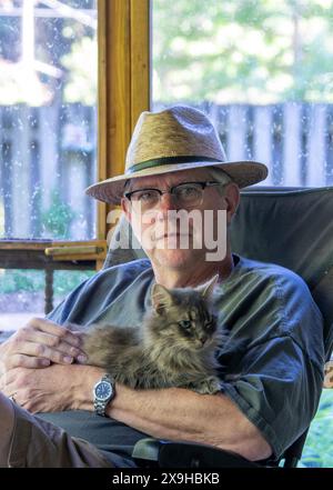 Homme plus âgé avec un chapeau de paille câlin avec son chat sur un porche moustiquaire Banque D'Images