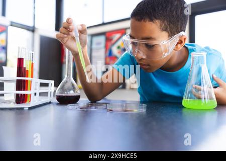 Biracial Boy participe à une expérience scientifique à l'école dans la salle de classe. Portant des lunettes de sécurité, il examine attentivement les réactions chimiques dans un tube à essai Banque D'Images