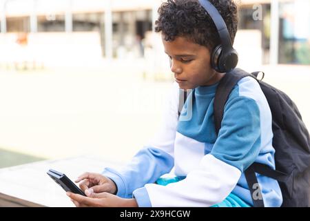 Biracial Boy aime la musique sur son téléphone à l'extérieur à l'école, avec un espace de copie Banque D'Images