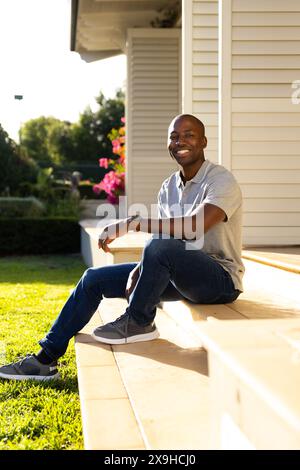 Jeune homme afro-américain assis sur des marches devant la maison, souriant à la caméra Banque D'Images