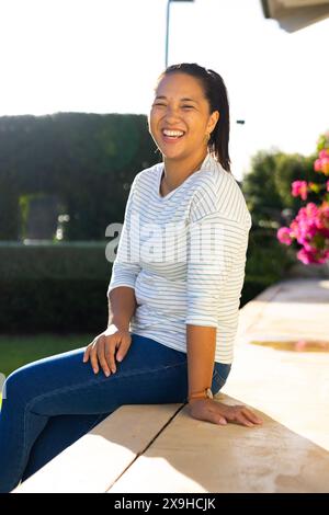 À l'extérieur, jeune femme biraciale souriant tout en étant assise sur le rebord en plein soleil Banque D'Images