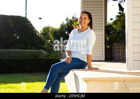 À l'extérieur, femme biraciale dans la trentaine assise sur les marches du porche, souriant chaleureusement Banque D'Images