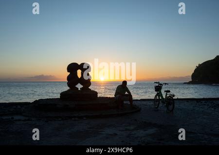 Aperçu de deux statues Tiki et d'un cycliste au coucher du soleil dans le petit village pittoresque d'Omoa, Fatu IVA, îles Marquises, Polynésie française Banque D'Images