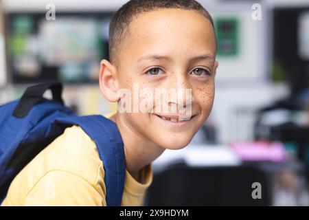 Garçon biracial avec des taches de rousseur souriant, portant une chemise jaune et un sac à dos dans une classe d'école Banque D'Images