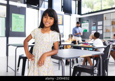 Fille biraciale avec de longs cheveux foncés se tient avec confiance dans une salle de classe d'école avec espace de copie Banque D'Images