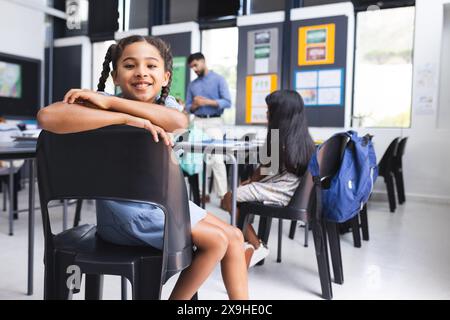 Fille biraciale avec des sourires de cheveux tressés, assise dans une salle de classe d'école avec un espace de copie Banque D'Images