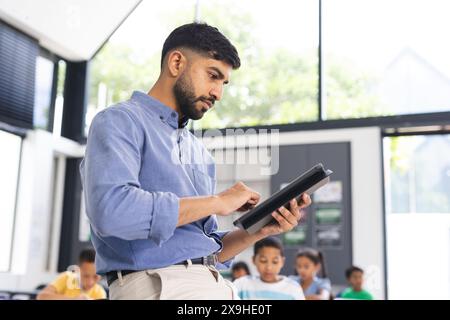 À l'école, un jeune professeur asiatique tenant une tablette se tient près des élèves dans la salle de classe Banque D'Images