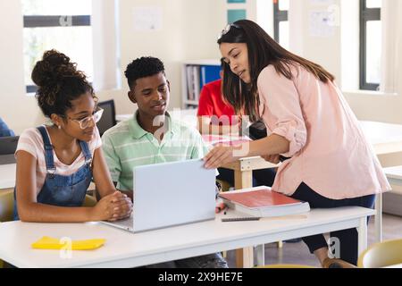 Jeune enseignante biraciale enseigne dans une salle de classe lumineuse au lycée. Elle aide des étudiants biraciaux adolescents avec un ordinateur portable, favorisant une colla Banque D'Images