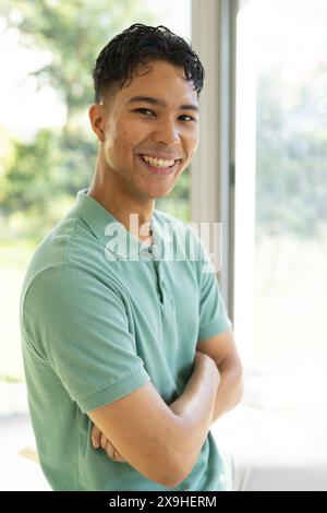 À la maison, un physiothérapeute masculin biracial sourit, les bras croisés dans une pièce lumineuse et aérée. Grandes fenêtres montrant la verdure à l'extérieur, créant un arrière-plan paisible Banque D'Images