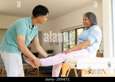 À la maison, physiothérapeute masculin biracial et patiente senior faisant des exercices de jambe. Ils sont dans une chambre lumineuse et confortable avec de grandes fenêtres et confortab Banque D'Images