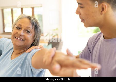 À la maison, physiothérapeute masculin biracial et patiente senior dans une pièce lumineuse. Grandes fenêtres et décor moderne créant un fond accueillant pour Banque D'Images