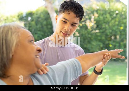 À la maison, physiothérapeute masculin biracial et patiente senior faisant des exercices de bras. Ils sont à l'extérieur dans un jardin, avec une végétation luxuriante en arrière-plan, Banque D'Images