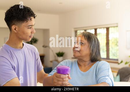 À la maison, le jeune physiothérapeute masculin biracial aide un patient féminin senior à exercer, en utilisant des haltères. Ils sont dans une salle lumineuse, bien éclairée avec grand Banque D'Images