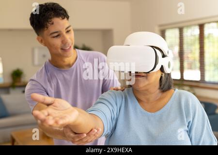 À la maison, physiothérapeute masculin biracial et patiente senior utilisant un casque VR. Ils sont dans une chambre lumineuse et moderne avec de grandes fenêtres et un confort Banque D'Images