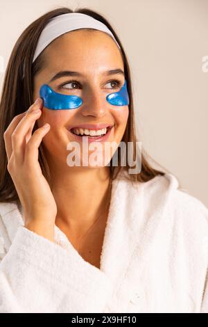 À la maison, une jeune femme caucasienne applique des patchs oculaires bleus, souriant et regardant de côté. Elle porte un peignoir blanc dans un environnement de spa à la maison lumineux et confortable Banque D'Images